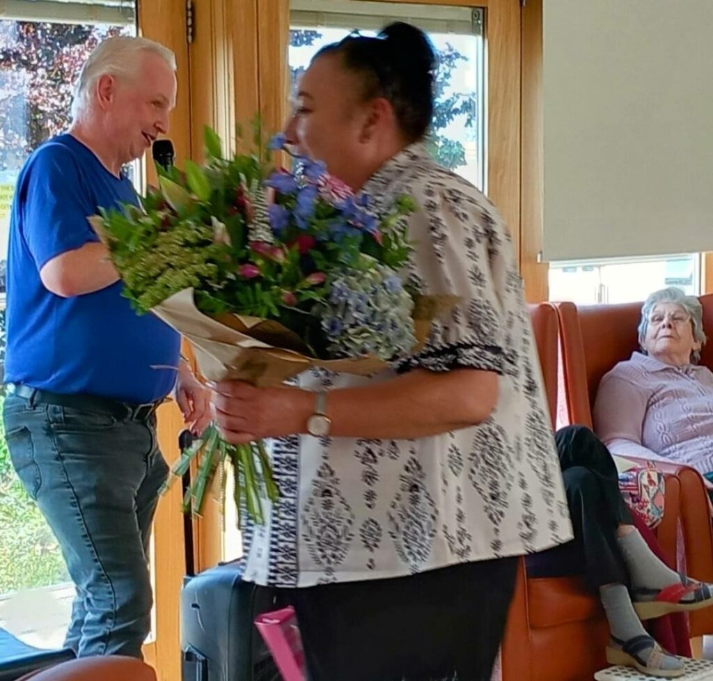 Sarah, recognised for her 28 years dedication, holds a bouquet of flowers as she walks past seated people in a bright room with large windows.