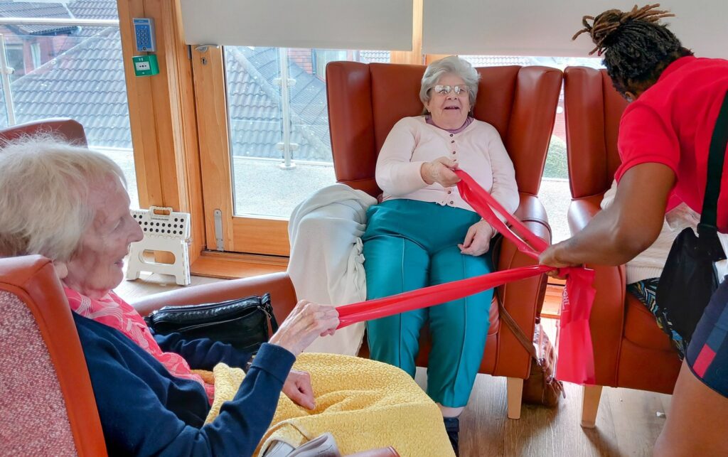 Two elderly women seated in armchairs take part in a lively exercise activity with a red resistance band, supported by a carer in a red shirt, setting an energetic tone for the start of August.