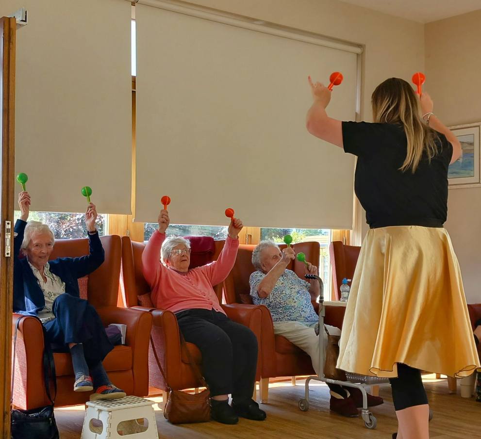 A group of elderly women seated in chairs raise maracas as a standing instructor leads an exercise or musical activity in a room with large windows.