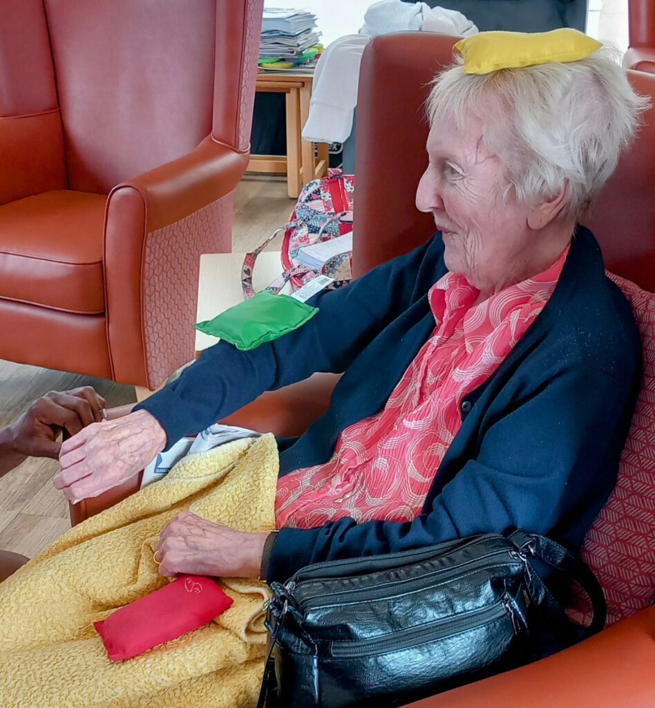 An elderly woman sits in a chair with a yellow cushion on her head, a green cushion on her arm, and a red cushion on her lap, holding hands with someone out of frame as the start of August brings warmth and comfort.