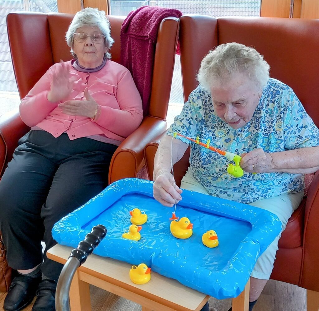 Two elderly women are seated in armchairs; one is playing a fishing game with toy ducks in a blue inflatable paddling pool, while the other claps her hands.