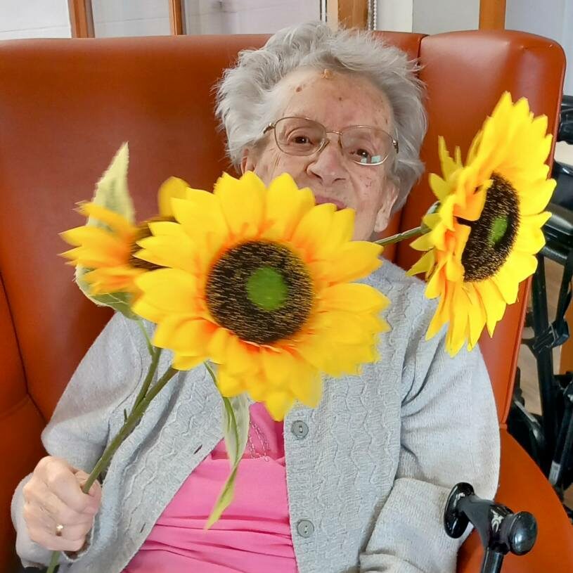 An elderly woman seated in an armchair holding two large artificial sunflowers, wearing spectacles, a pink top, a grey cardigan, and a floral skirt.