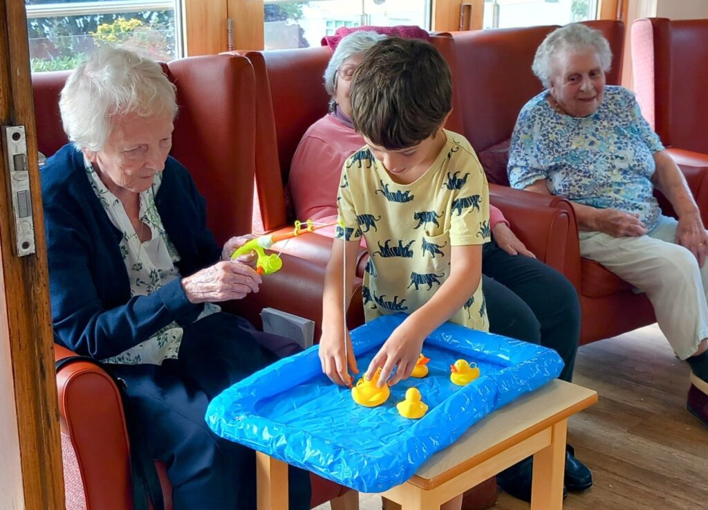 An elderly woman and a boy play a fishing game with toy ducks in a blue inflatable paddling pool, while two other elderly people watch from red armchairs in a bright room.