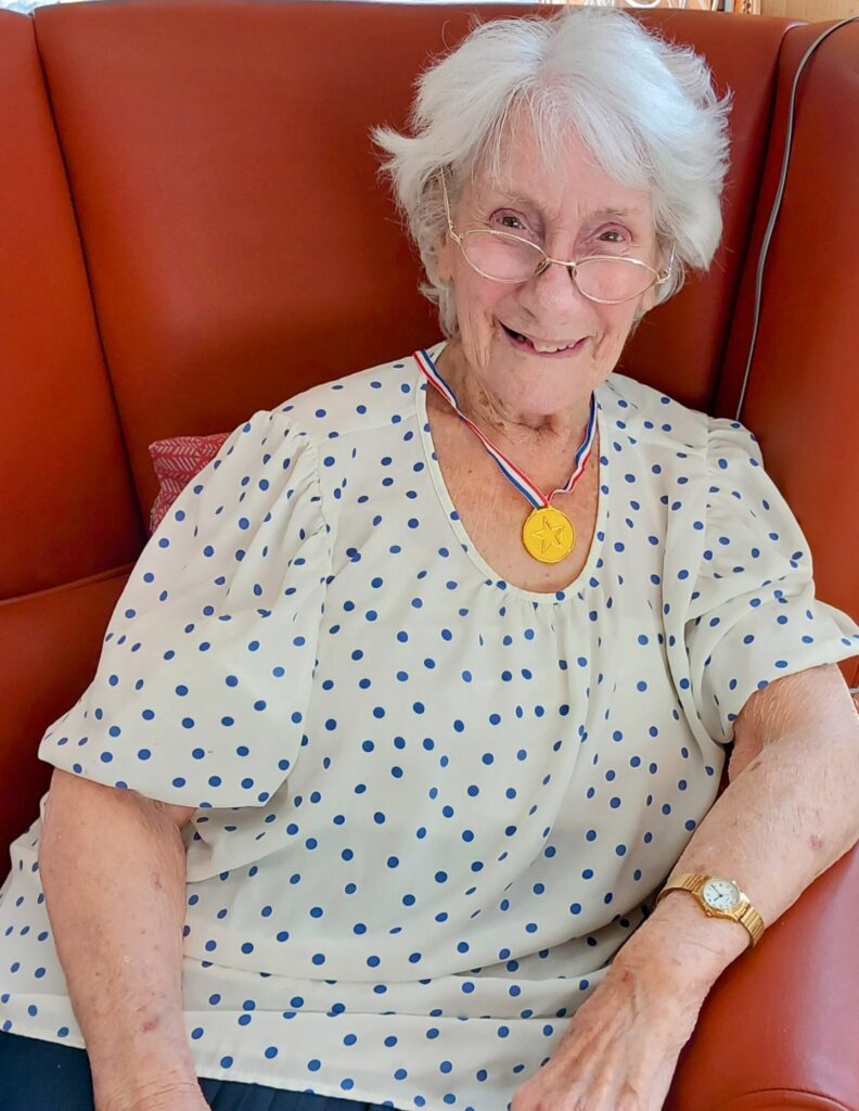 Elderly woman with white hair and spectacles, dressed in a polka dot blouse and gold medal, seated on a red chair and smiling at the camera, capturing a true Golden Moment.
