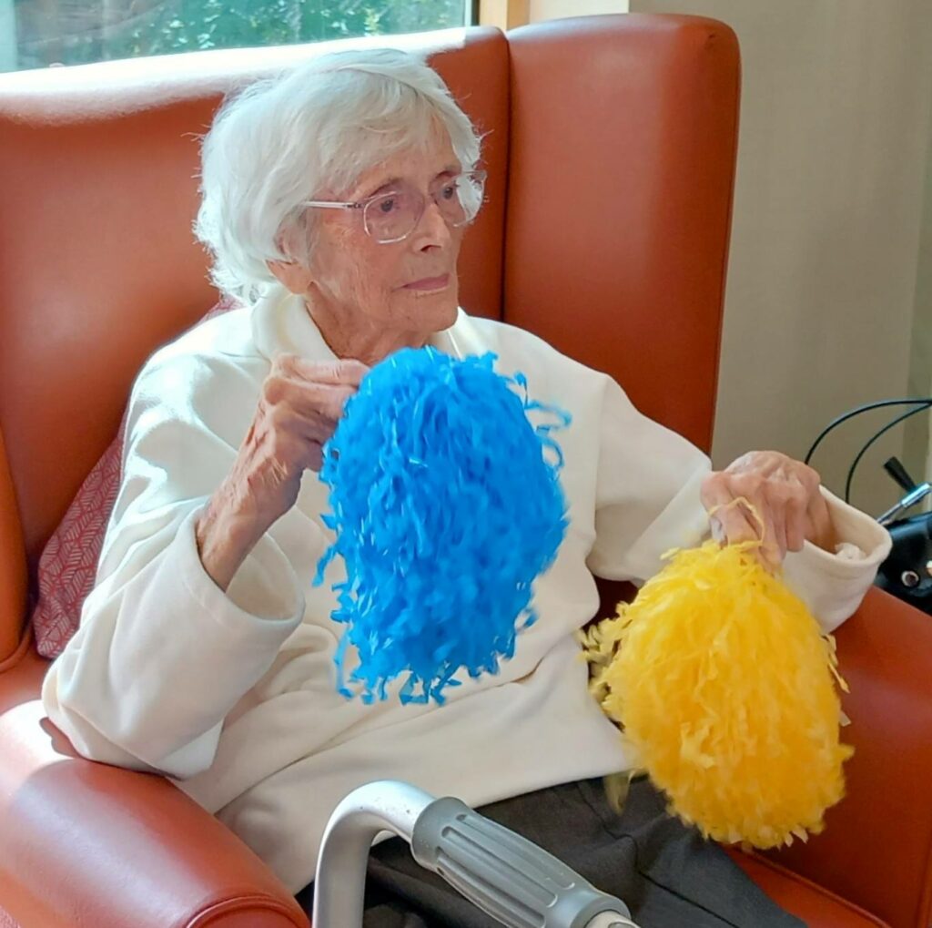 An elderly woman with white hair sits in an orange chair, holding blue and yellow pom-poms. She wears glasses and a white jumper, capturing a golden moment of fun.