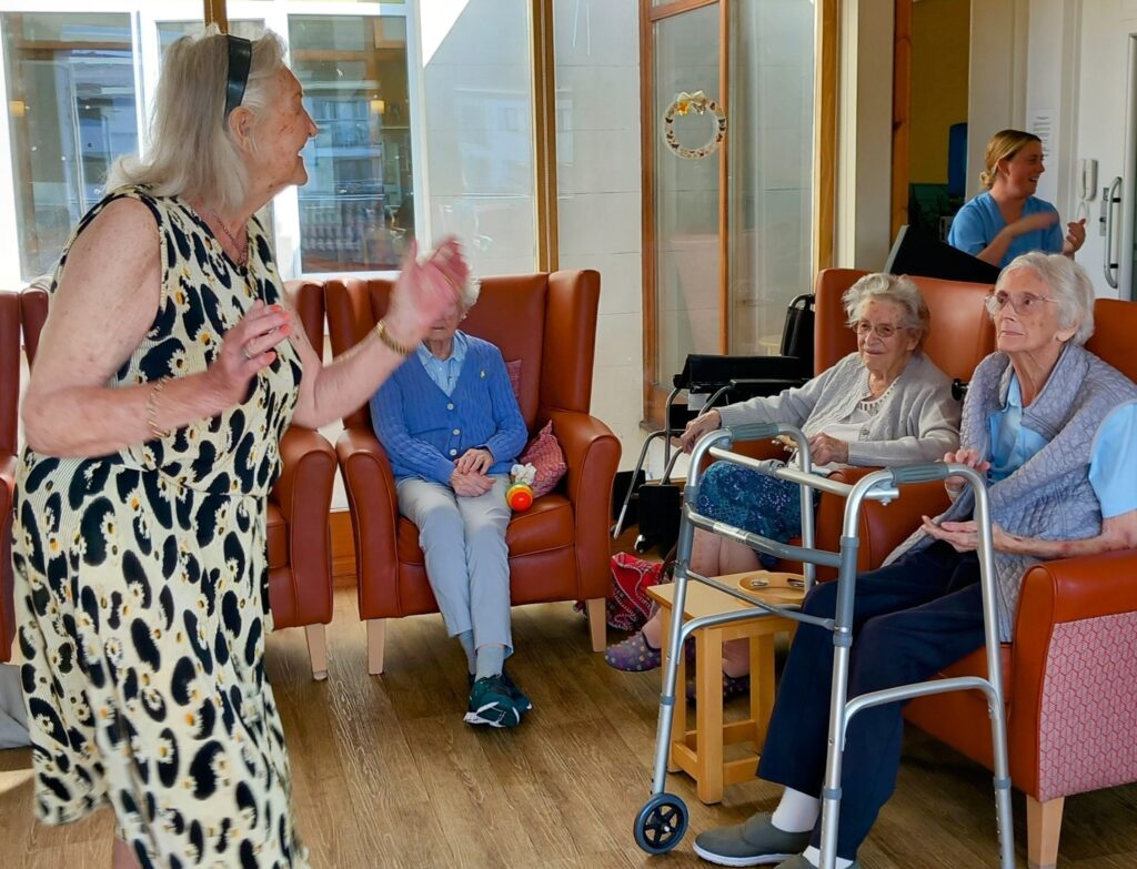 A woman dances before three seated elderly ladies, two with walking frames, in a brightly lit common room—creating a cheerful afternoon for everyone as a staff member stands by the door in the background.