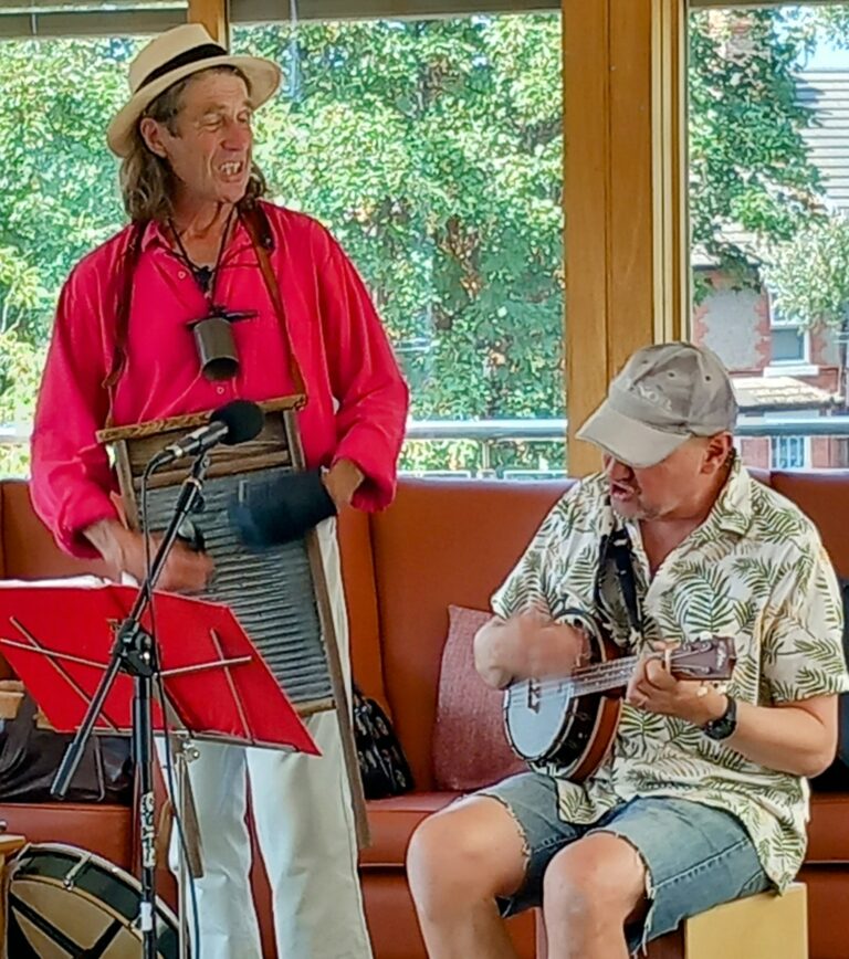 Marko and Andrew enjoy a fun afternoon indoors—Marko stands playing a washboard in his hat, while Andrew sits strumming a banjo. Trees and houses can be seen through the window behind them.