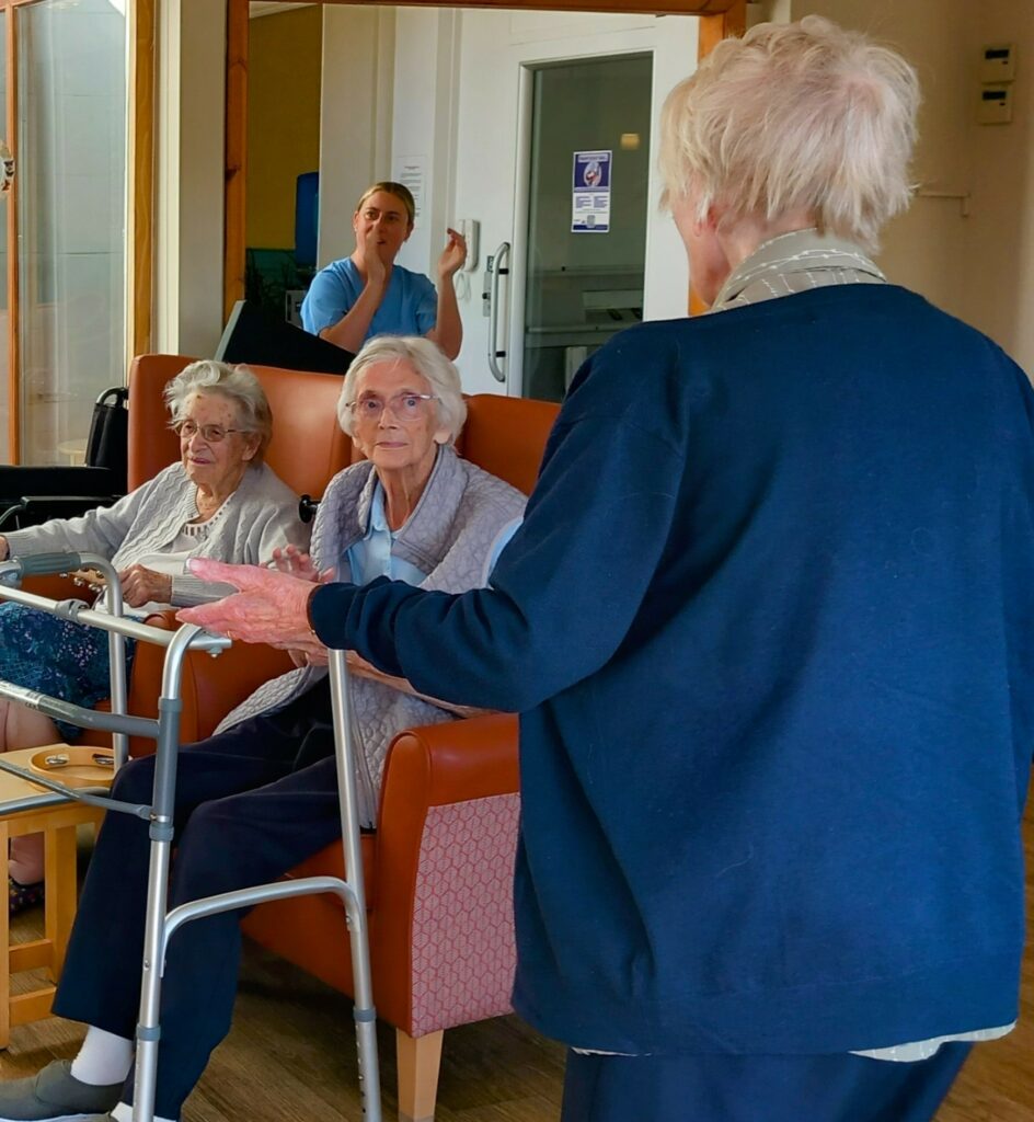 Three elderly women have an enjoyable afternoon chatting in a care home lounge, with one using a walking frame. Marko, a staff member, is visible in the background near the door.