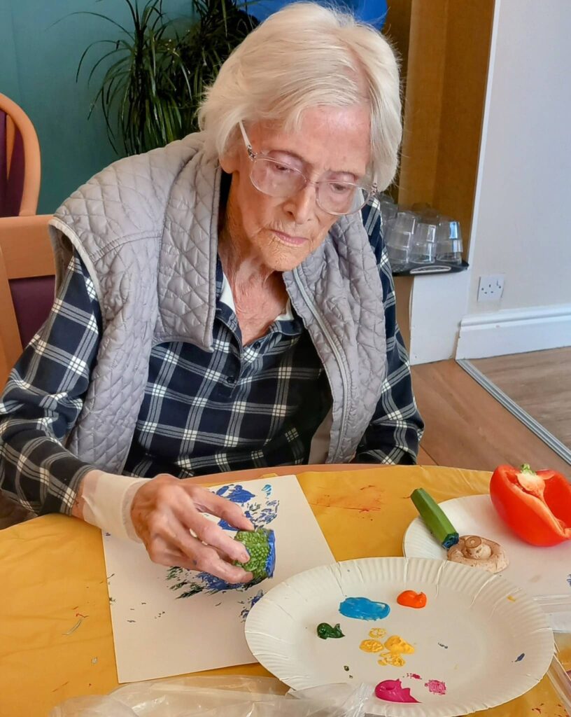 Celia enjoys an August Crafternoon, pressing broccoli dipped in colourful paint onto paper at a table, surrounded by vibrant paint splodges and fresh vegetables.
