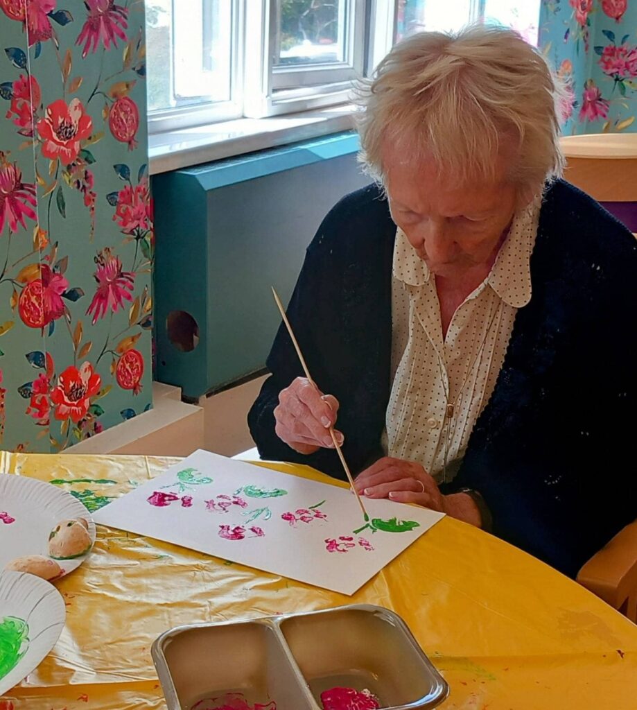 Celia, an elderly woman, sits at a table covered with a yellow cloth during an August Crafternoon, painting colourful flowers on paper using her brush and paint supplies.