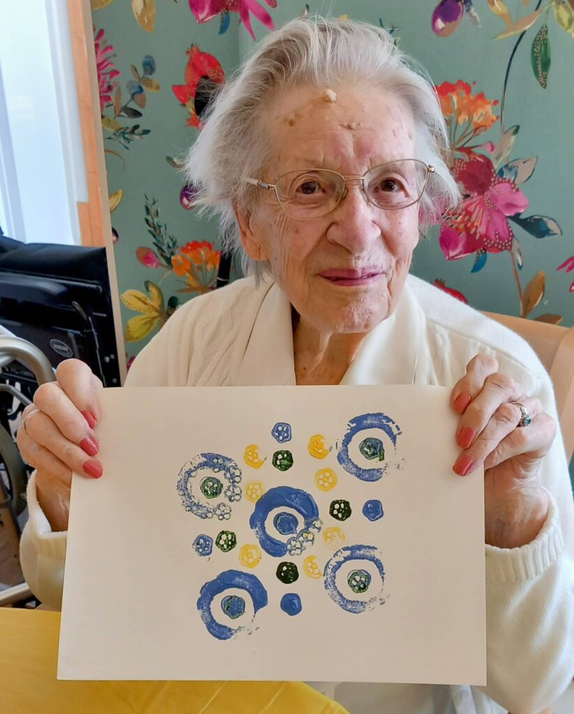 Celia, an elderly woman with glasses, holds up a sheet of paper showing a colourful abstract artwork with blue and yellow circular patterns at August Crafternoon.