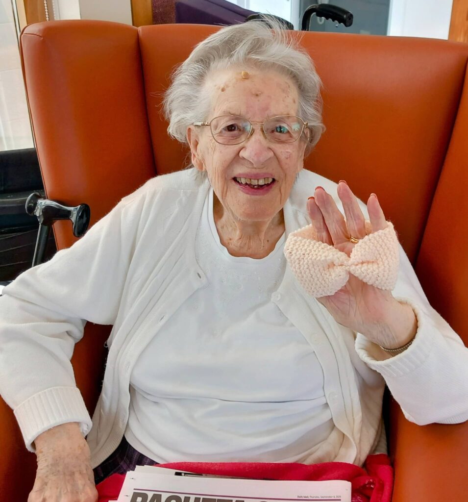 Elderly woman seated in an orange chair, smiling and holding up a knitted bow at Knit and Natter. She is wearing glasses, a white blouse, and a white cardigan. A newspaper lies on her lap.