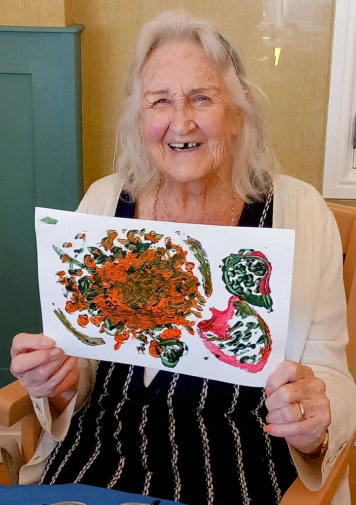 Celia, an elderly woman, smiles as she holds up a colourful hand-painted artwork featuring orange and green shapes, possibly flowers or fruit, made during an August Crafternoon indoors.