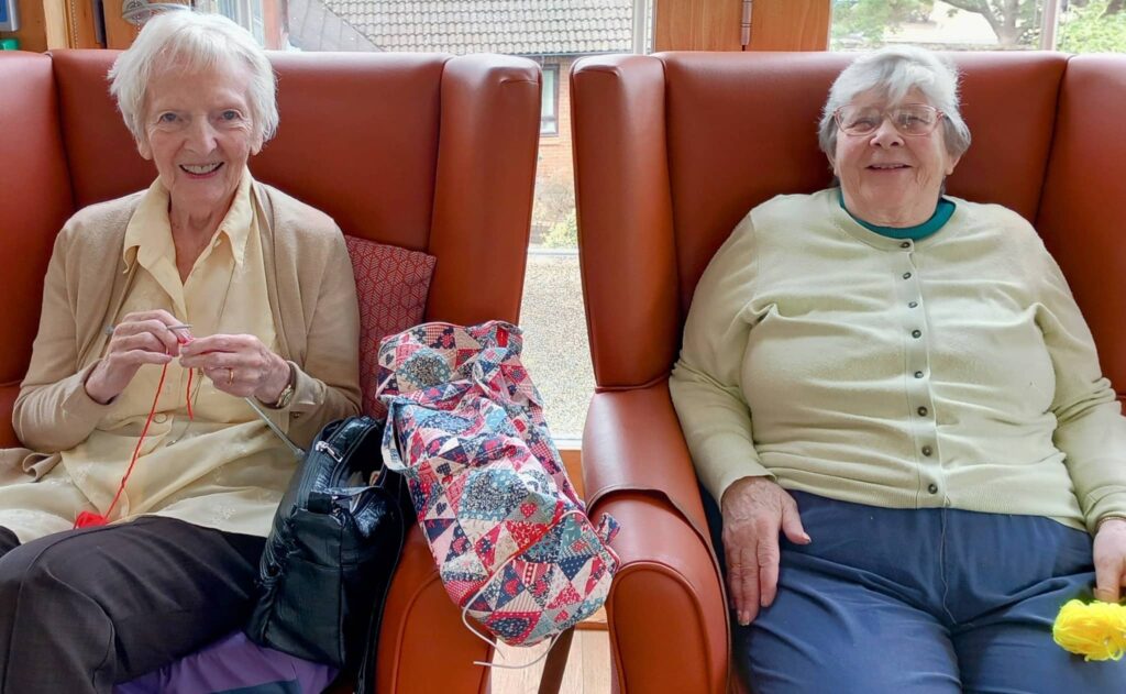 Two elderly women enjoy their later years, smiling as they take part in a Knit and Natter session—one knits with red wool while a patterned bag sits between their orange chairs.