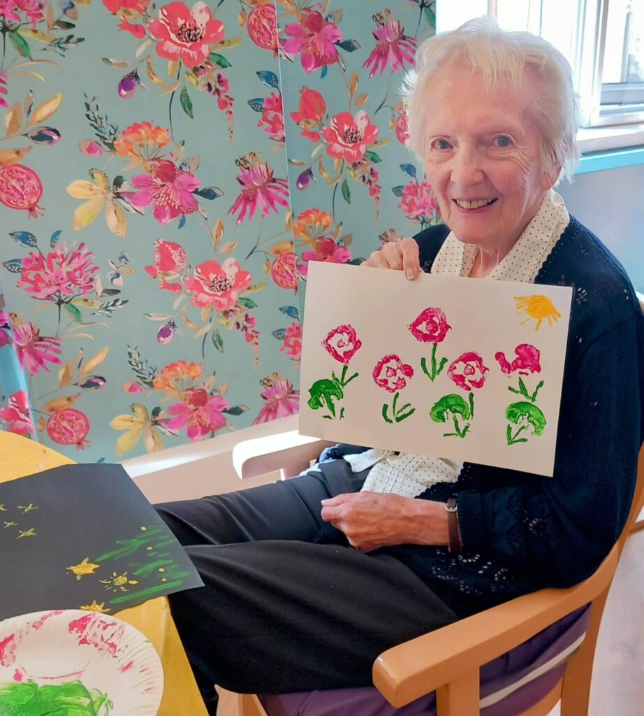 Celia, smiling at the camera, sits in front of a floral curtain and proudly shows her painting from a Colourful August Crafternoon. Art supplies are scattered across the table before her.