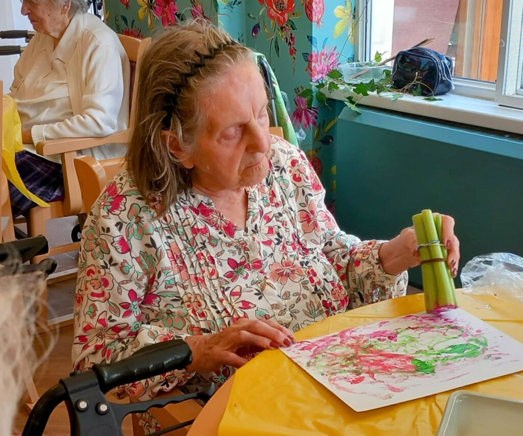 During the August Crafternoon, Celia, an elderly woman, sits at a table stamping a sheet of paper with celery sticks to make colourful artwork in a bright, decorated room.