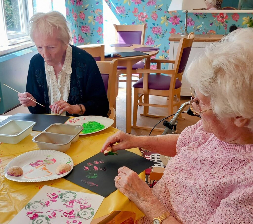 Two elderly women, including Celia, enjoy a colourful August Crafternoon, painting on black paper with brushes amidst art supplies in a brightly lit, floral-decorated room.