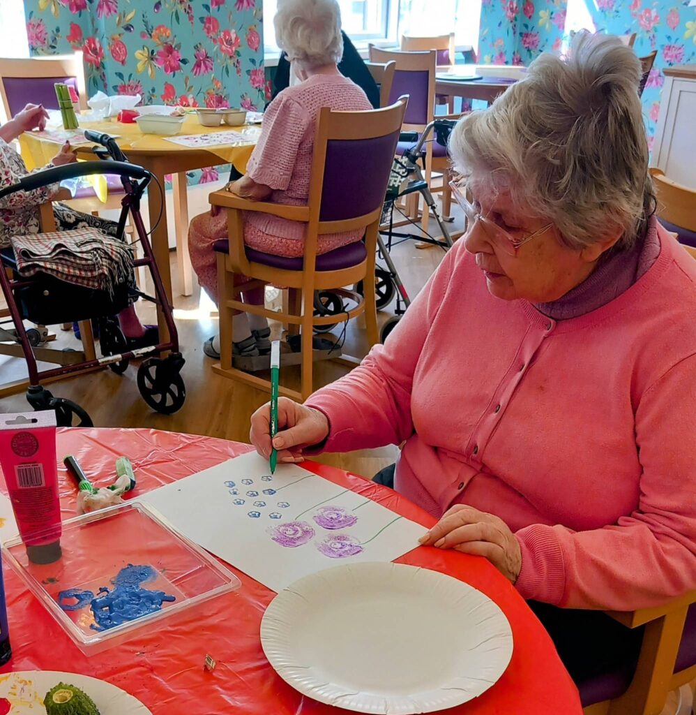 Celia, an elderly woman, sits at a table drawing with felt-tip pens on paper during an August Crafternoon, while other pensioners enjoy the colourful, brightly decorated room in the background.