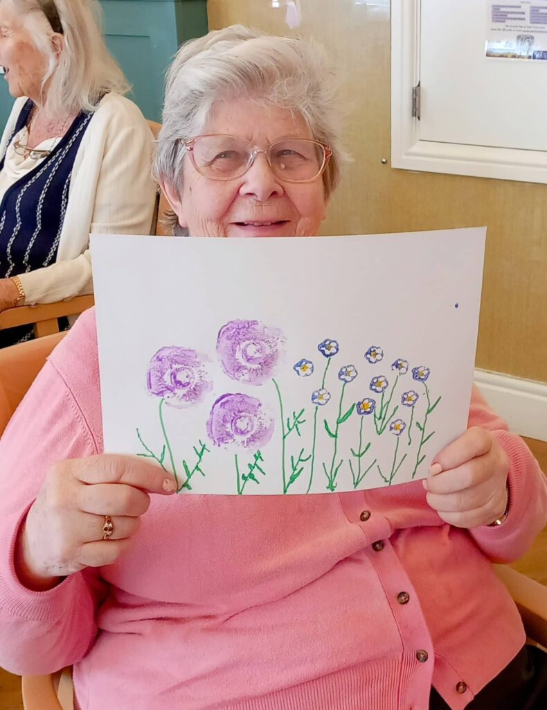 Celia, an elderly woman wearing spectacles, holds up a drawing of purple and yellow flowers she created during a Colourful August Crafternoon while seated indoors.