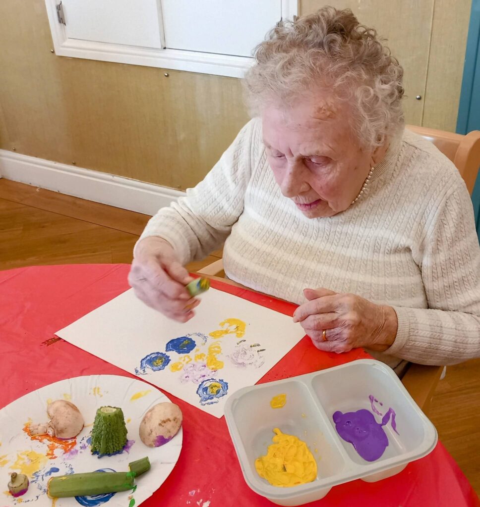 Celia enjoys a colourful August Crafternoon as she sits at a table, using vegetables and paint to make prints on paper. Paint and vegetables are set out on a plate and in a divided tray in front of her.