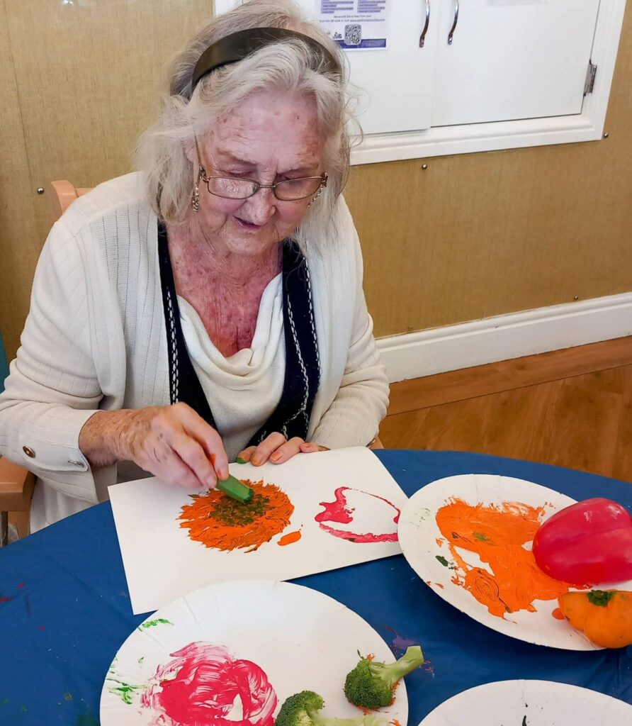 During an August Crafternoon, Celia uses broccoli and a pepper to apply colourful paint to paper at a table, with paint and vegetables on plates.