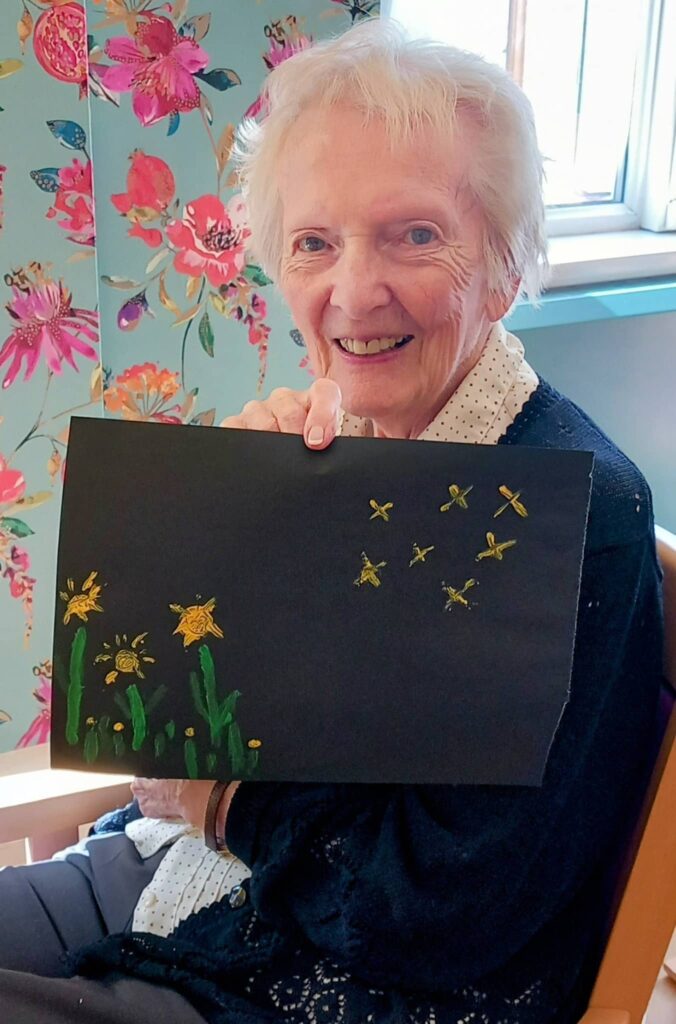 Celia enjoys a colourful August Crafternoon indoors, holding up a black sheet of paper decorated with yellow and green drawings of flowers and bees.
