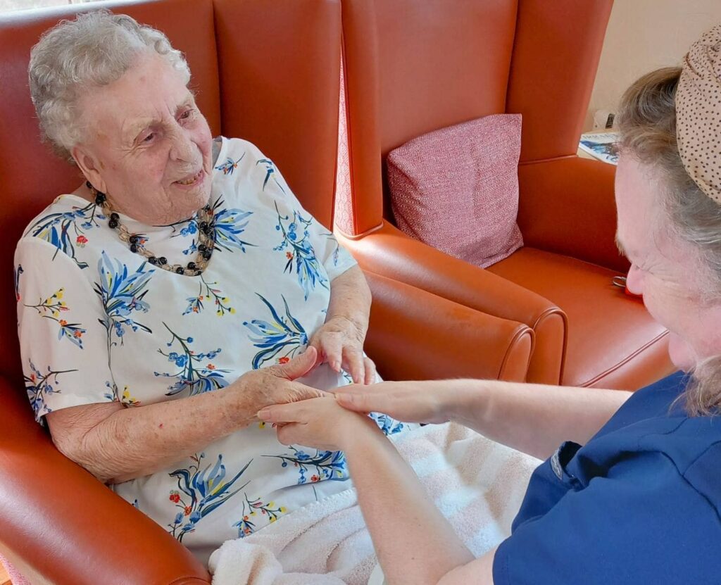 An elderly woman sits in an armchair smiling as another woman, perhaps a carer, gently massages her hands—reflecting the warmth and comfort of Pamper Afternoons at Abbeyfield in later life.