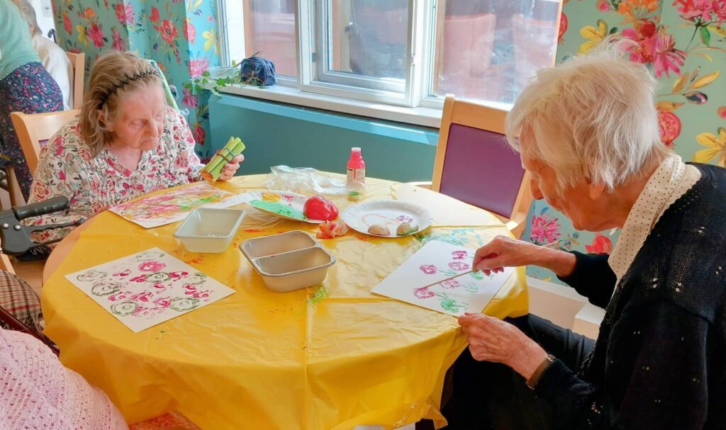 Two elderly women sit at a table covered with colourful art supplies, painting on paper in a brightly decorated room with floral wallpaper during an August Crafternoon.