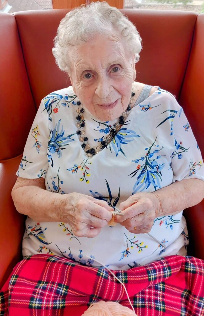 Elderly woman with short grey hair sits in a red chair, wrapped in a red checked blanket, enjoying a creative moment as she knits with peach-coloured yarn—an inspiring scene of later life at its finest.