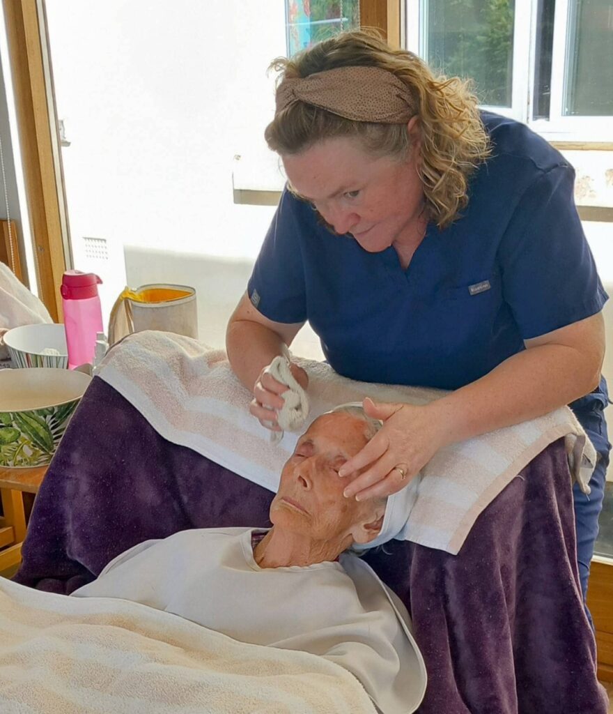 A woman in navy scrubs cleans the face of an elderly person lying back with a towel around their head in a well-lit room during Pamper Afternoons at Abbeyfield.