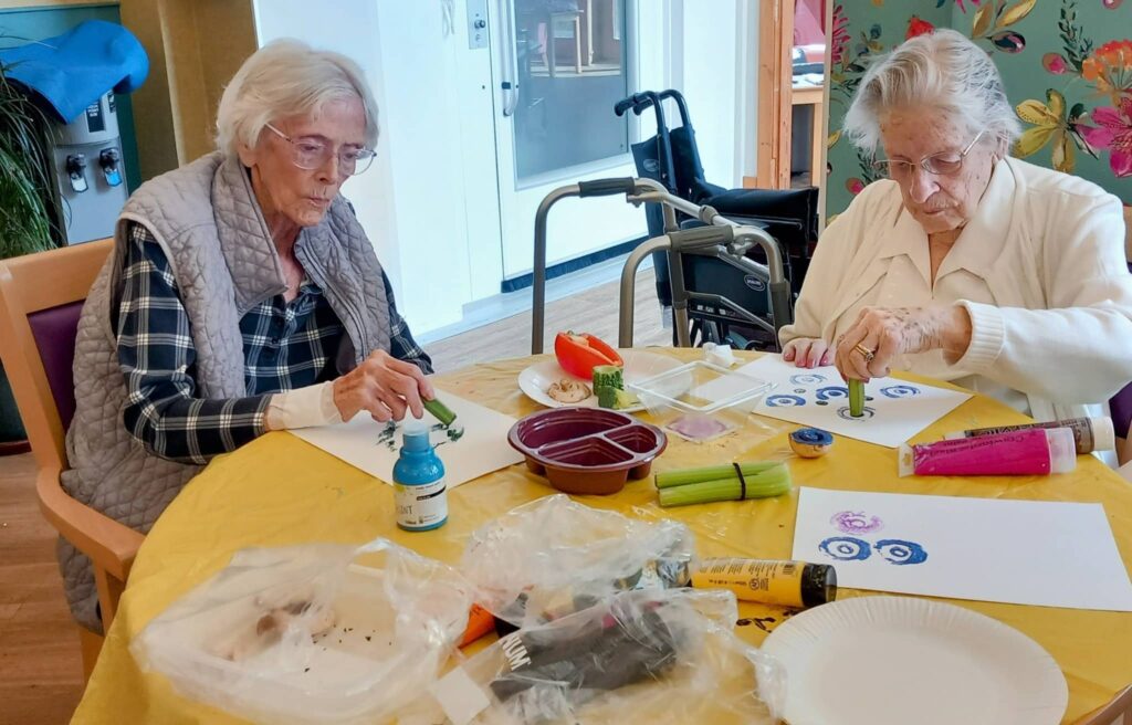Celia and a friend sit at a table in a brightly lit room, using vegetables and paint to create artwork during a Colourful Crafternoon.