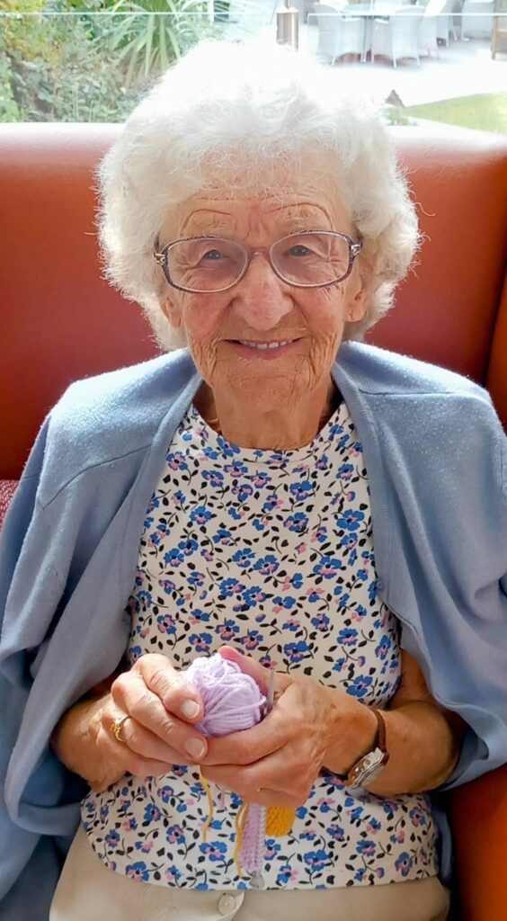 Elderly woman with white hair, glasses, and a light blue shawl sits on a red chair, smiling and holding a small knitted item—demonstrating her creativity at a Knit and Natter gathering in later life.