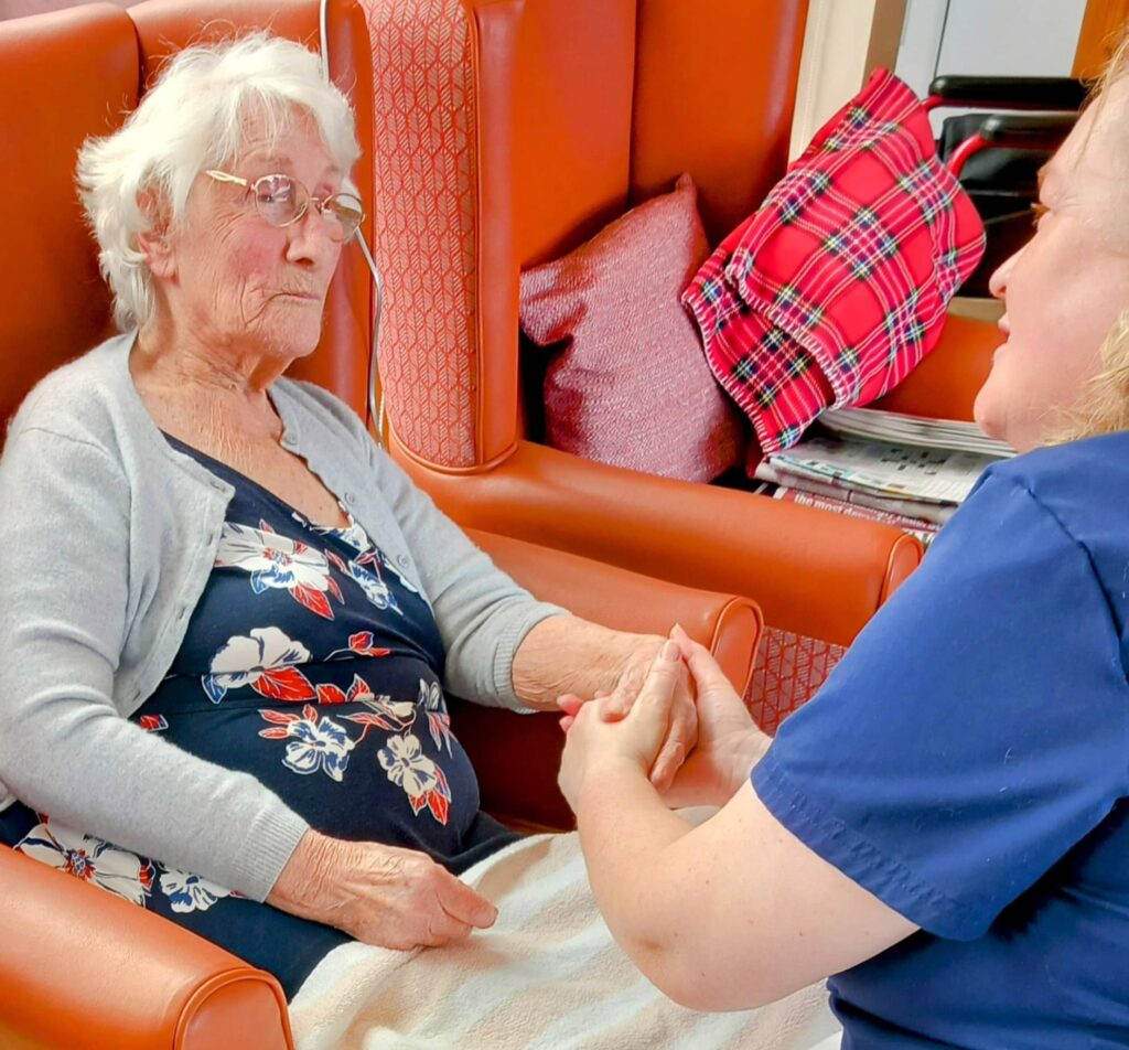 An elderly woman sits in an armchair, holding hands with a carer and sharing a warm moment during Abbeyfield’s supportive Pamper Afternoons.