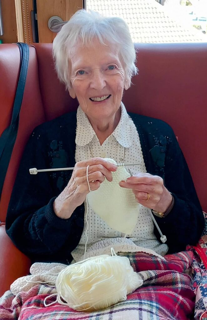 An elderly woman with white hair sits in a red armchair, smiling as she knits with white wool and needles—a cosy scene of later life as she enjoys her Knit and Natter group. A checked blanket covers her lap.
