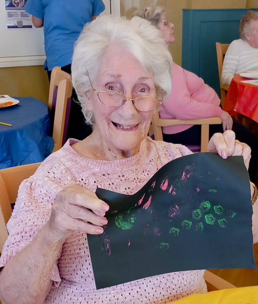 Elderly woman wearing glasses smiles as she holds up a piece of colourful abstract artwork made during an August Crafternoon in a communal setting.