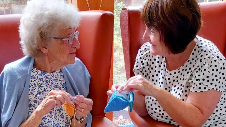An elderly woman and a younger woman sit in armchairs, sharing their creativity as they knit and have a natter together, smiling warmly at each other in later life.