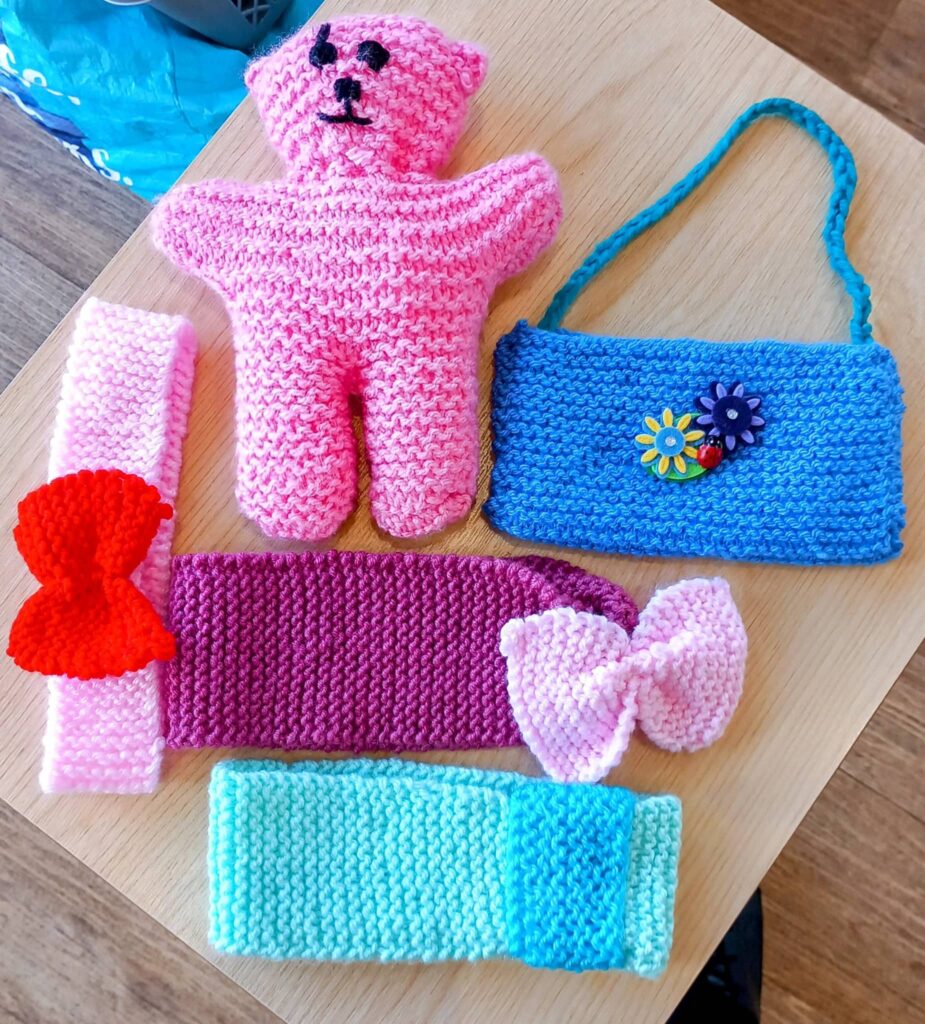 Demonstrating later-life creativity, these Knit and Natter makes feature a pink teddy bear, blue purse with flower buttons, colourful headbands, and two delightful knitted bows arranged on a wooden table.