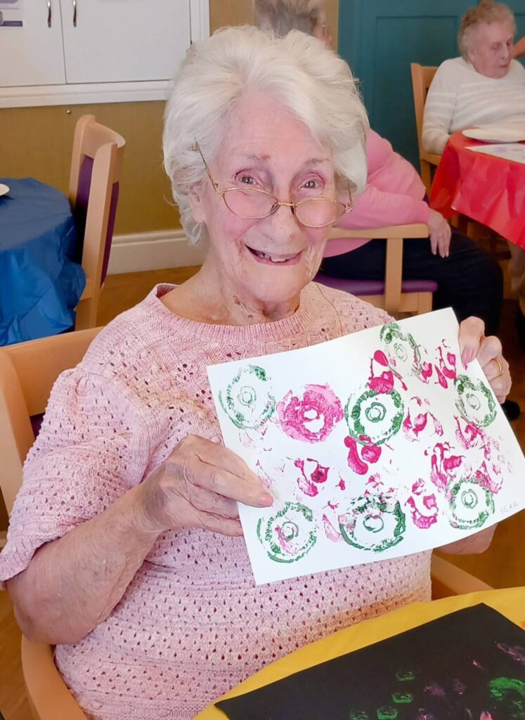 Celia, wearing glasses and a pink jumper, smiles as she proudly shows off her colourful circular artwork from the Colourful August Crafternoon in the communal room.