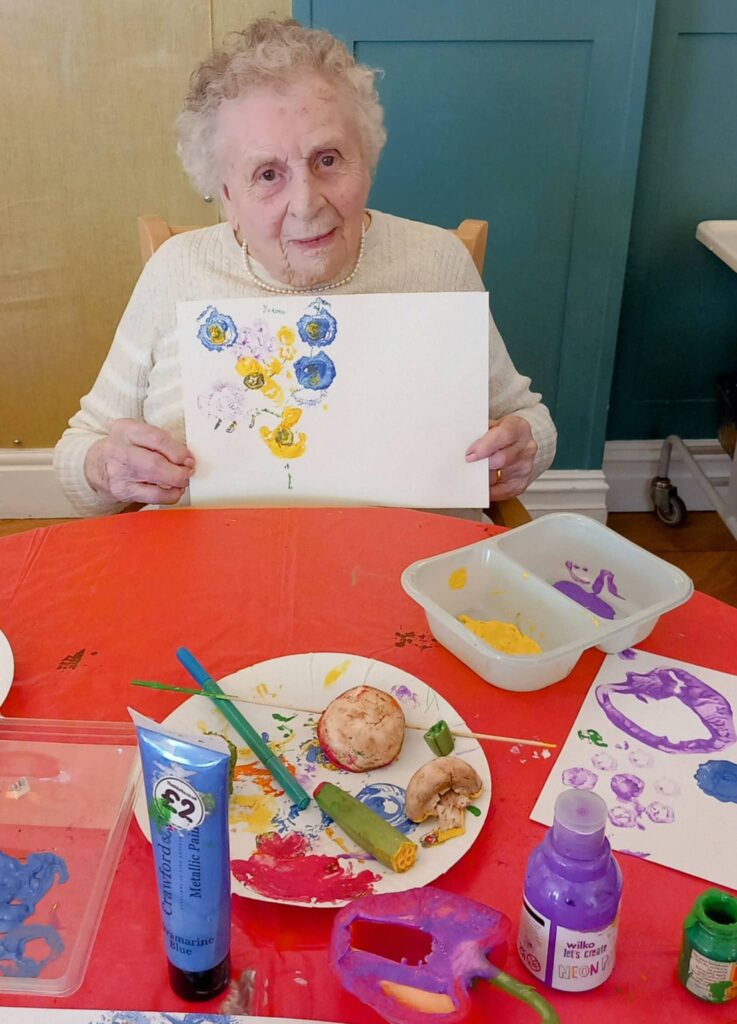 Celia, an elderly woman, sits at a table during August Crafternoon, proudly holding up her colourful flower painting amongst painting supplies, vegetables, paint and brushes.