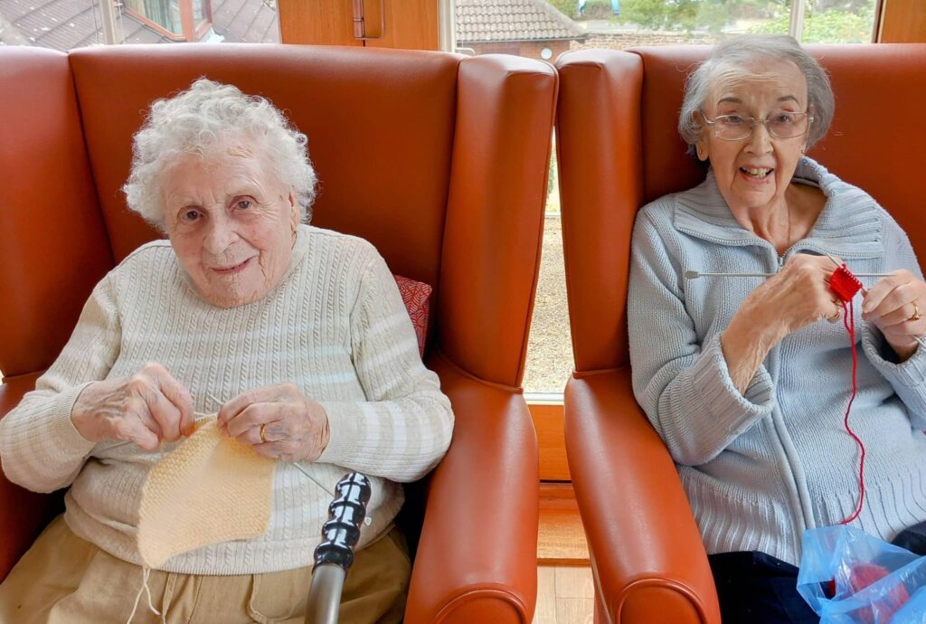 Two elderly women sit in red chairs by a window, both knitting and smiling at the camera, sharing creativity and conversation in later life as part of a cheerful Knit and Natter gathering.