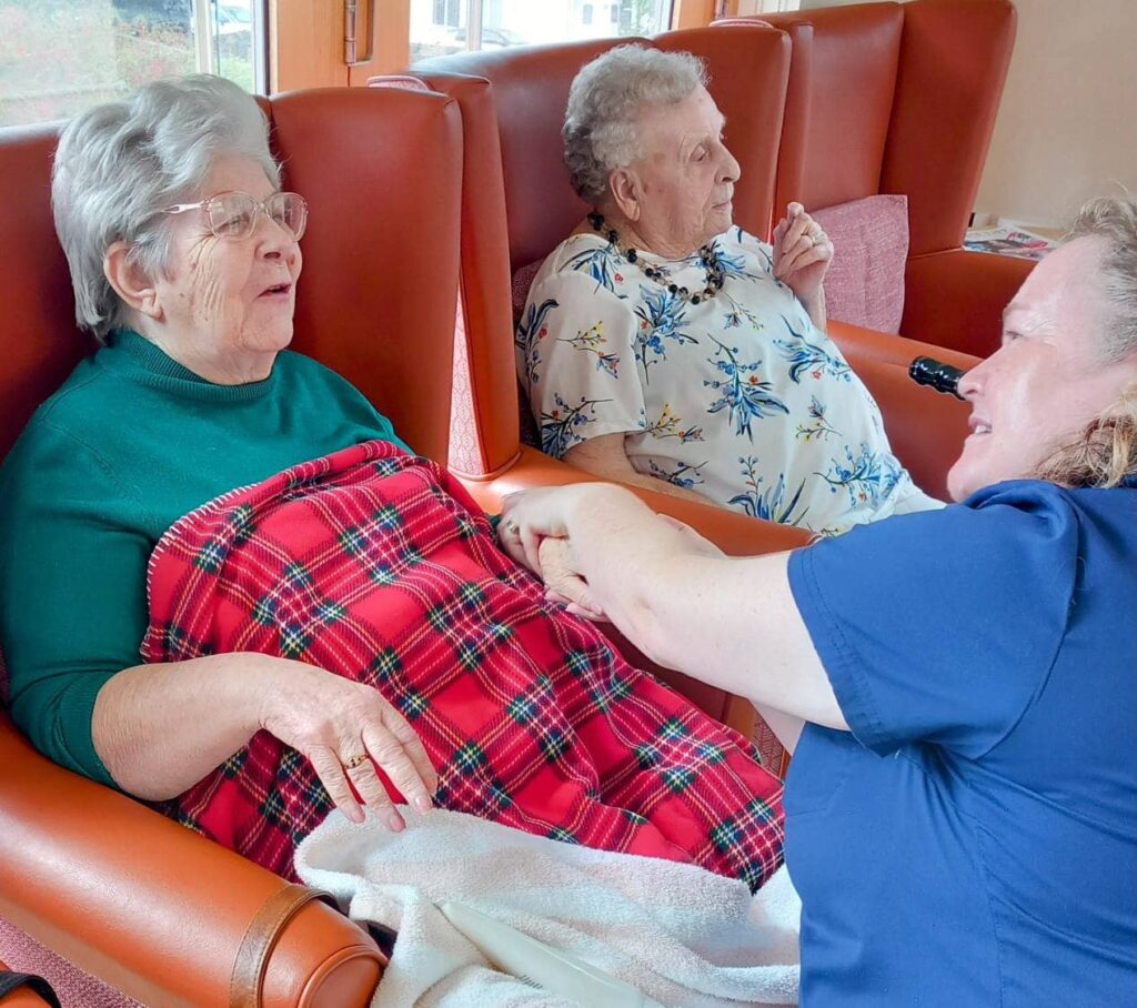 Two elderly women sit in armchairs, one draped with a red tartan blanket, as a carer in an Abbeyfield uniform kneels beside them and gently holds one lady’s hands, creating a comforting and relaxed atmosphere in later life.