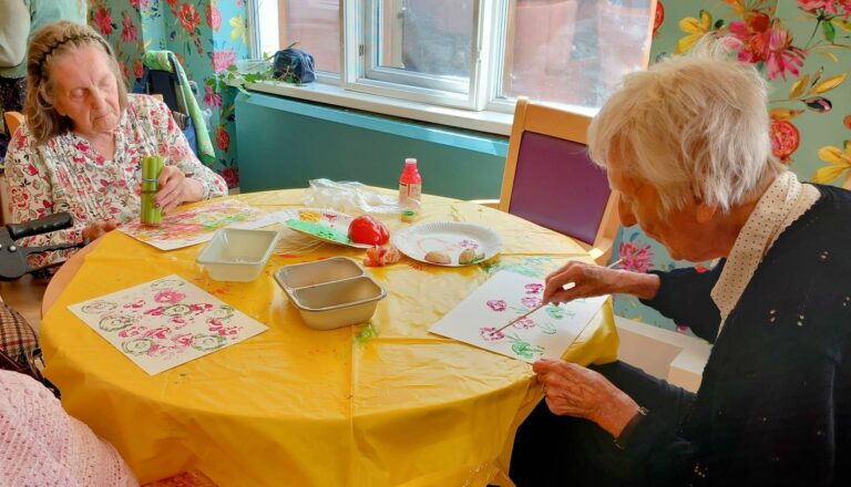 Celia and another elderly woman sit at a round table draped with a yellow cloth, enjoying a colourful August crafternoon as they create vibrant art prints on paper using paint and vegetables.
