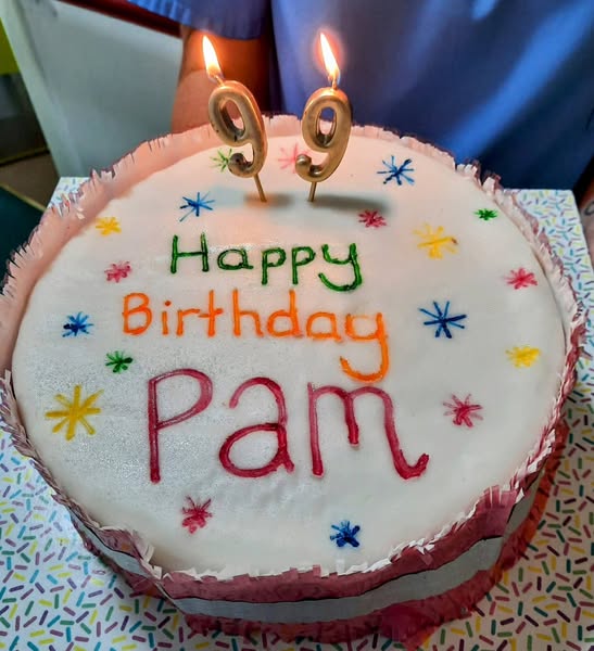 A birthday cake with 'Happy Birthday Pam' written on it and two lit candles shaped as the number 99.