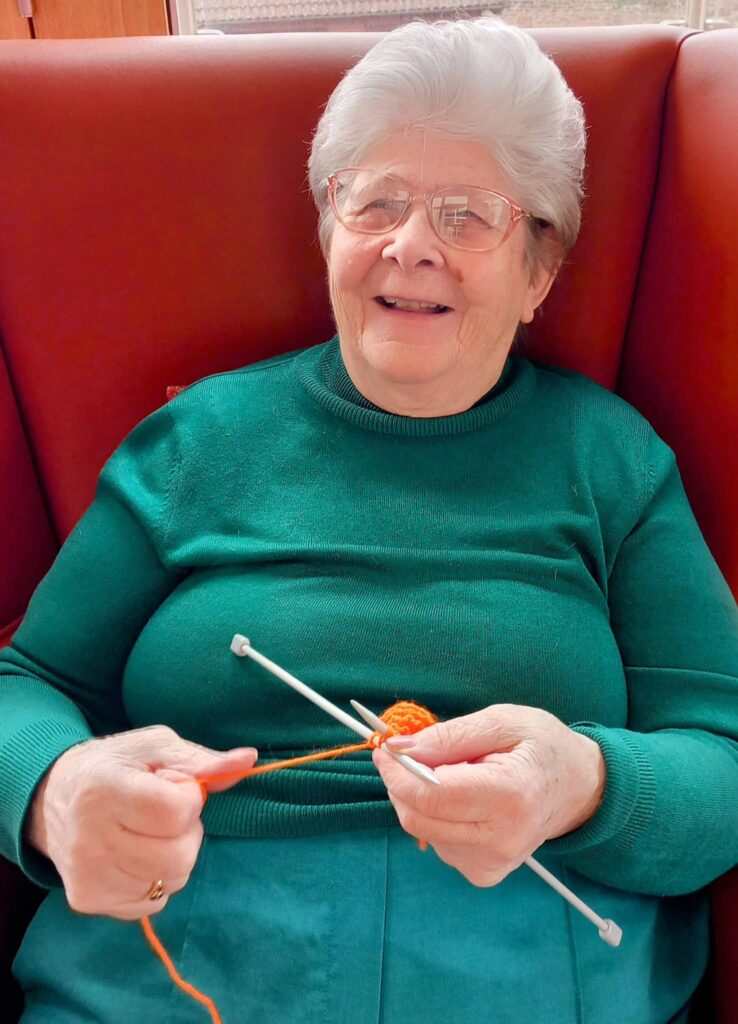 An elderly woman with grey hair and glasses sits on a red chair, smiling as she knits with orange wool and needles—her creativity shining through during a relaxing Knit and Natter session in later life.
