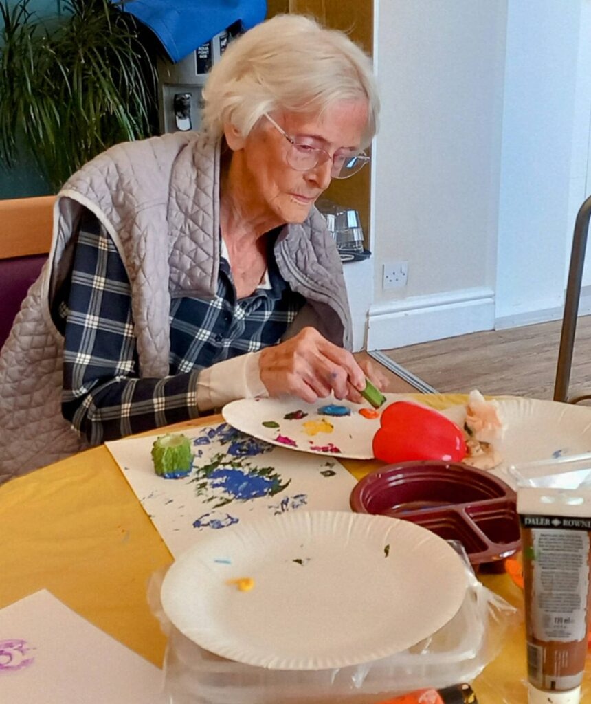 Celia, an elderly woman, sits at a table painting on paper with colourful paints, surrounded by art supplies and paper plates during an August Crafternoon.