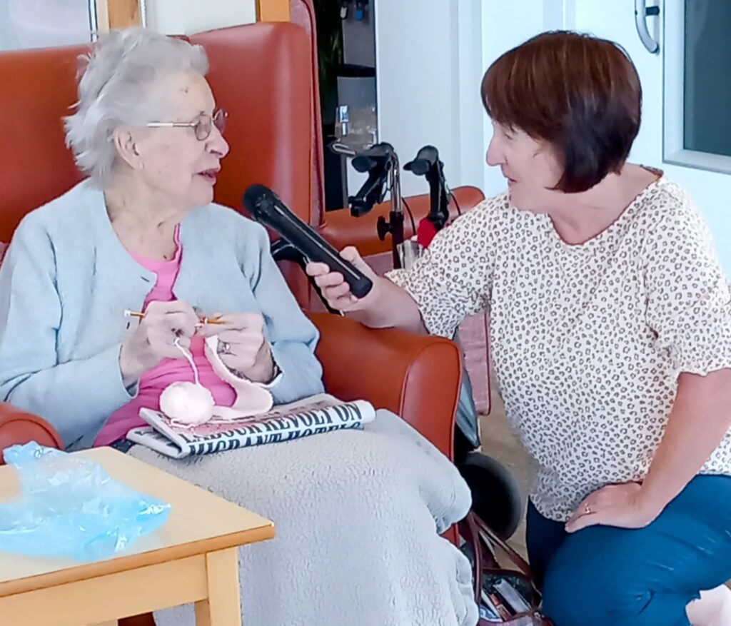 An elderly woman embraces creativity in later life as she knits during a Knit and Natter session, while another woman kneels next to her, holding a microphone for an interview.