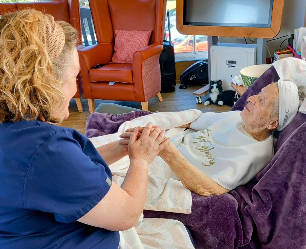 A carer holds the hands of an elderly woman lying in a recliner, covered with towels and a blanket during Pamper Afternoons at Abbeyfield, bringing comfort and warmth to later life in a brightly lit room.