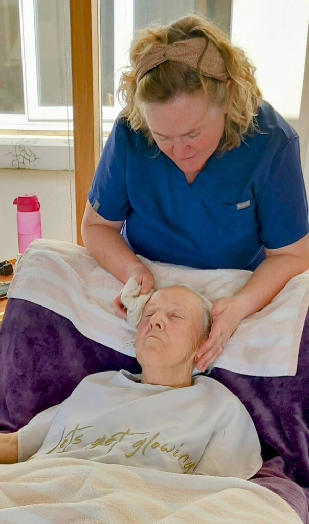 A carer gently wipes the face of an elderly woman resting in a chair, covered with a towel and blanket, enjoying Pamper Afternoons in the sunlit Abbeyfield room.