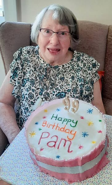 An elderly woman sits on a sofa and smiles behind a decorated birthday cake with "Happy Birthday Pam" and a number 99 candle on top.
