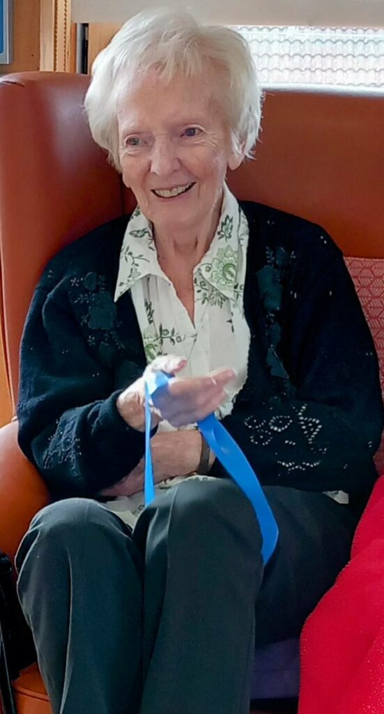 Elderly woman with short white hair sitting in an armchair, smiling and holding a blue ribbon or strap in her left hand.